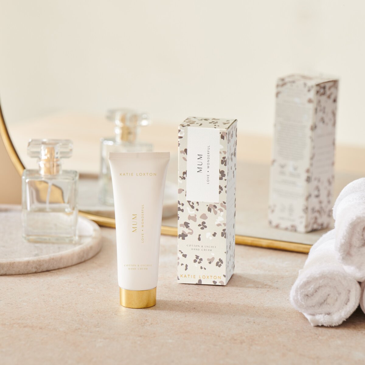 Tube of Mum handcream sitting on a dressing table next to its floral box, a mirror and some rolled hand towels.