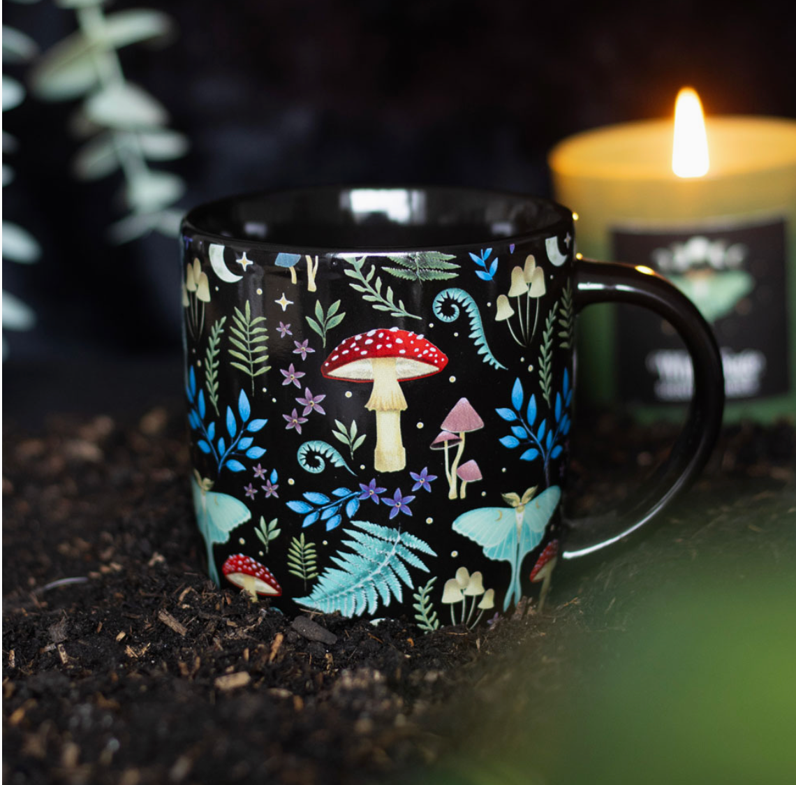 Black mug with a 'mystical forest' print with toadstools, moths, various foliage a star and moon. There is a lit candle in the background
