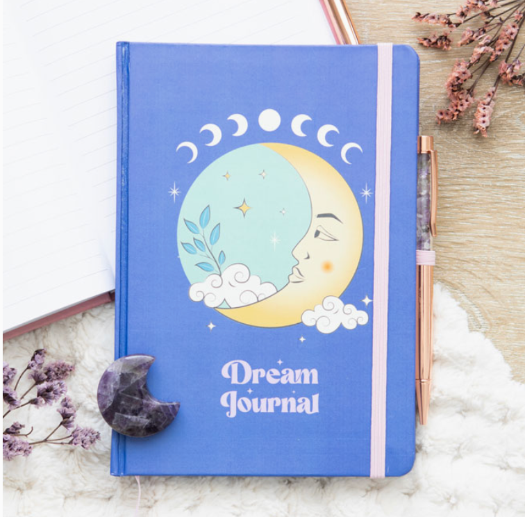 blue dream jounral notebook with celestial moon design, pink elastic strap and rose gold pen filled with amethyst chips it sits on a desk with dried flowers and a fluffy white mat