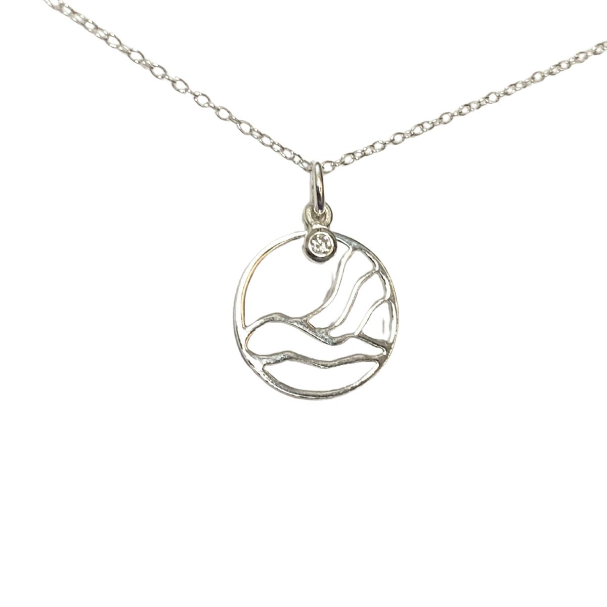 close up of northern lights inpired pendant. The pendant is round, with wavy lines deonting the land and 3 wavy lines representing the northern lights. There is a small round clear crystal at the top of the pendant. It hangs on a silver chain.
