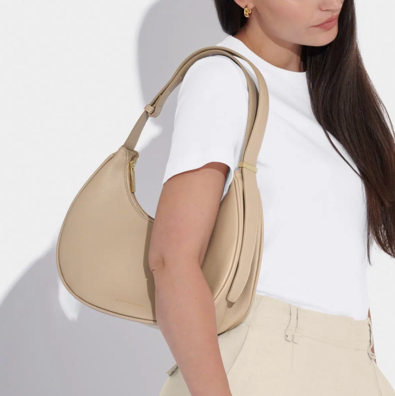 model with long brown hair wearing a white t-shirt and taupe trousers with the light taupe scoop style handbag on her right shoulder.