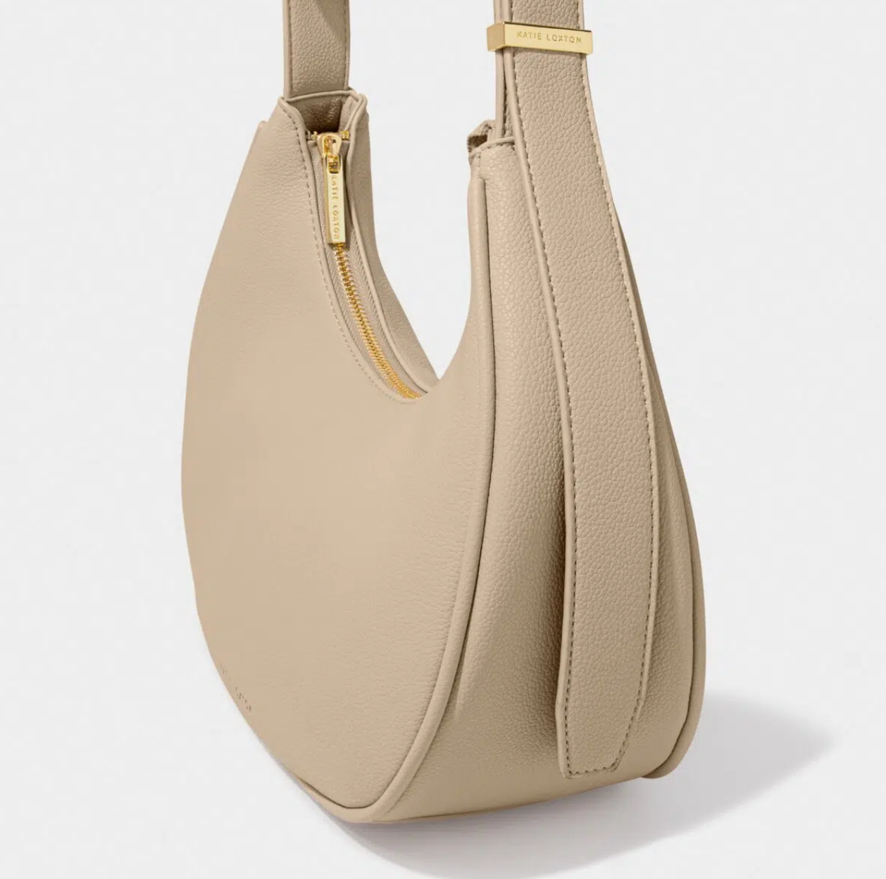 side view of the light taupe scoop handbag showing the gold zip and gold strap hardware, which both say Katie Loxton. The bag sits against a white background