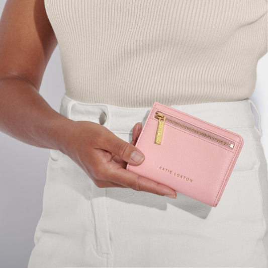 Model wearing a taupe top and white trousers holding the Katie Loxton Jayde purse in cloud pink. The purse has a gold zip and katie loxton in gold lettering