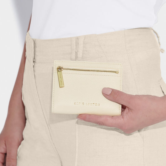 Model wearing a white top and taupe trousers holding the Katie Loxton  Jayde purse in Ecru. The pruse is a small rectangle with a gold zip.