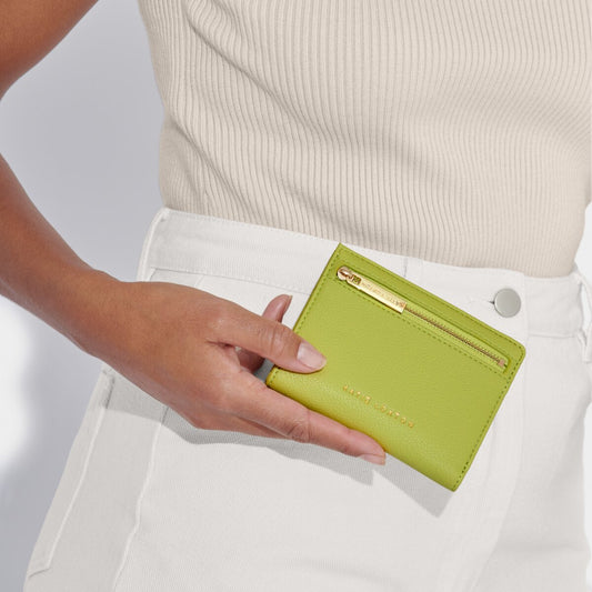 model wearing a cream top and white jeans holding the katie loxton Jayde purse in lime green. the purse is a small rectangle with a gold zipper