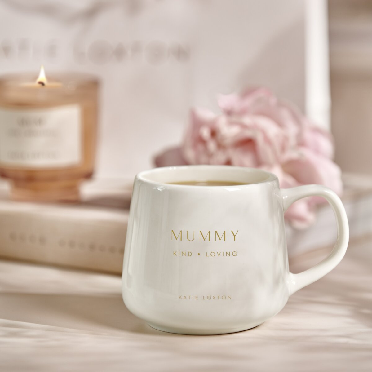 White mug with 'mummy' 'kind' and 'loving' stamped in gold on the front. A burning candle , flowers and a book in the background